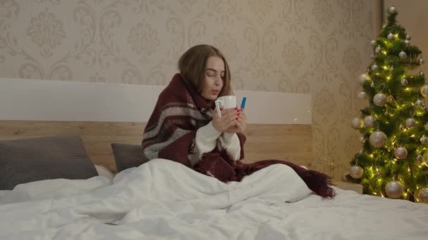 一个年轻的女人在圣诞节和新年假期病倒了 她正坐在家里的卧室里 毛毯铺满 手里拿着一杯饮料和药品 — 图库视频影像