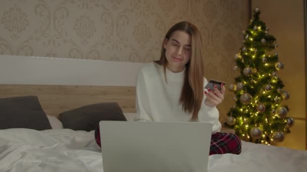 若い女性はオンラインで家族のための休日のギフトを購入し 銀行カードで支払います ベッドルームでパジャマの女性がオンラインショッピングをし 宅配を待っています 冬休みコンセプト — ストック動画