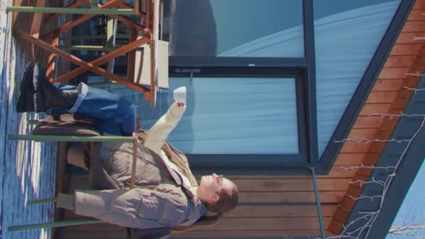テラスに座っている女性は パノラマウィンドウ付きの彼女のプライベートハウスのホットコーヒー垂直ビデオプライベートハウステラスを飲みます この瞬間は 民家での国内至福の本質を象徴しています — ストック動画