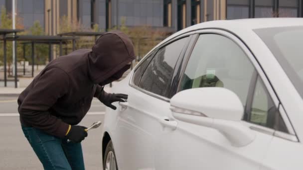 駐車場では 手のキーを握った泥棒が白い車の窓を揺さぶり 周囲の喧騒の中で広い日光の中で自動車の盗難の大胆な行為を開始します — ストック動画