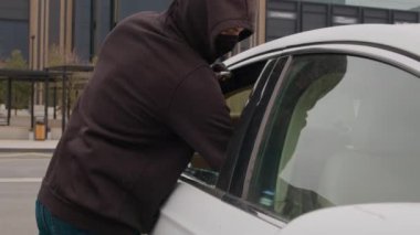 Siyah kapüşonlu bir hırsız, alışveriş merkezinde park etmiş bir arabanın kırık camından bilgisayar çalar.