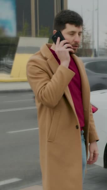 ベージュのコートの男は ストリートの垂直ビデオで電話会話で話しています ベージュのコートはコートで話される各単語のスタイリッシュな男が付いているマンフレームの上に優雅に引出します注意を時を超えない方法を指示します — ストック動画
