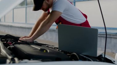 Mekanik arızaları tamir etmek için dizüstü bilgisayarı kullanarak kaputun altındaki aracı tamirci tamir ediyor. Kaput altındaki oto tamircisi teknoloji servis hizmeti kullanarak kaput altında çalışır