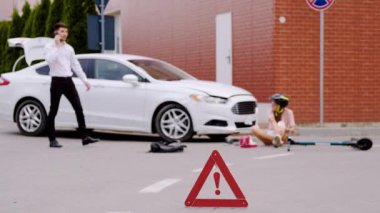 Araba-elektrikli scooter çarpışması kırmızı uyarı işareti trafik kazasına işaret ediyor. Adam yaralı kadın scooter sürücüsüne yardım etmek için acil servise telefon ediyor.