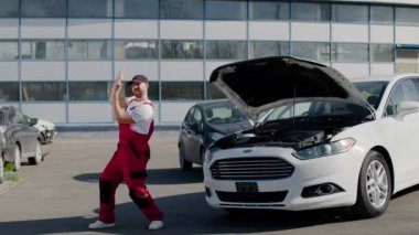 Erkek araba tamircisi otomobil tamirhanesinde arabanın yanında neşe içinde dans ediyor araba bakım merkezleri arabanın yanında neşeli bir şekilde dans ediyor. Tamirci otomobil servisinin arka planında mutlulukla dans ediyor