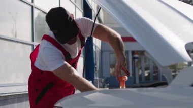 Araba servisindeki çalışma süreci. Kırmızı iş üniforması giymiş oto tamircisi arabanın kaputunun altında ingiliz anahtarıyla otomobil parçaları monte ediyor. Dışarıda bir araba servisinin önünde çalışan bir tamirci tamir ediyor.
