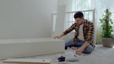 Genç adam mobilyaları monte ederek gelecekteki çocukların odasını hazırlıyor. Bebek için parlak ve minimalist bir oda. Babam yeni doğan için beşiği talimat kitabının yardımıyla birleştiriyor.