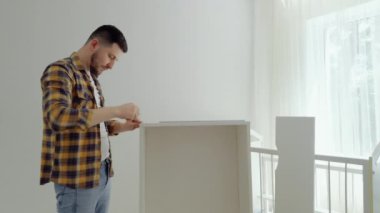 Mutlu adam çocuk odası için mobilyaları topluyor ve kameraya samimi bir şekilde gülümsüyor. Genç baba vidaları sıkıyor ve bebek karyolasını monte ediyor. Yeni doğan için mobilyaları hazırlıyorum.