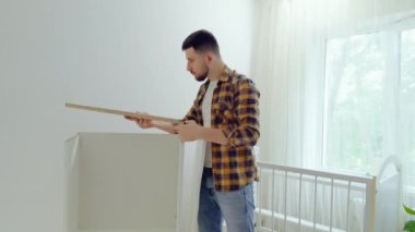 Genç adam çocuk odası için beyaz ahşap mobilya monte ediyor. Genç baba, bebek odasında gardırop yapmak için odun topluyor. Yeni doğan için mobilyaları hazırlıyorum.