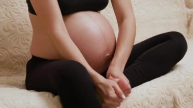 Hamile bir kadın evdeki kanepede otururken şişmiş bacağını ovalıyor. Tanımlanamayan anne, ayak bileğine masaj yapıyor ve hamilelik sırasında bacak krampları geçiriyor.