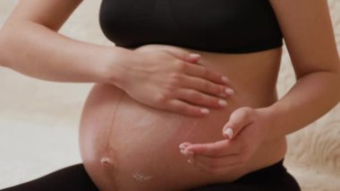 Hamile bir kadın son üç ay içinde karnına krem sürerek çatlakları önlüyor. Geniş göbekli hamile bir kadının, göbeğindeki kuru deriyi nemlendirip masaj yaptığı yakın plan görüntüsü.