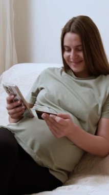 Genç hamile kadın kanepeye yaslanıyor ve akıllı telefondan kredi kartı bilgilerini giriyor. Gülümseyen kadın çevrimiçi alışverişe çıkıyor ve bebek için kıyafet alıyor.