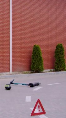 Devrilmiş elektrikli scooter ve kask kaldırımda kaza uyarısı levhasının yanında duruyor. Sahne çarpışmanın ciddi sonuçlarını gösteriyor. Kazadan sonra