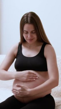 Gülümseyen hamile kadın masaj hareketleriyle çıplak karnına losyon sürüyor. Genç bir kadın annelik yolculuğu sırasında cilt bakımı prosedürü uygular.