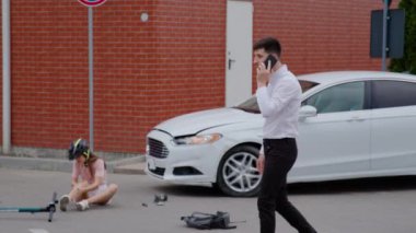 İş adamı kaza yapmış arabanın yanında acil bir telefon görüşmesi yapıyor. Miğferli kadın yerde oturup acıdan titreyen yaralı bacağını tutuyor. Emniyet Müdürü ve Polis Çağrısı