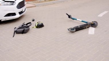 Hasarlı elektrikli scooter ve kask kazadan sonra çamurluğu ezilmiş beyaz arabanın yanında asfaltta yatıyor. Sahne kaza sonrasını gösteriyor. Son çarpışma belirtileri