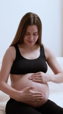 Gülümseyen hamile kadın kanepeye oturarak nemlendirici krem sürüyor. Siyah elbiseli neşeli genç bayan cilt bakımı rutinine başladı.