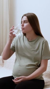 Genç hamile kadın bardaktan takviye su alıyor. Müstakbel anne koltukta otururken sağlığıyla ilgilenir.