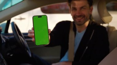 Arabadaki gülümseyen adam akıllı telefondaki araba paylaşım uygulamasını gösteriyor. Adam kısa süreli araç kiralama hizmetinden memnun olduğunu ifade ediyor.