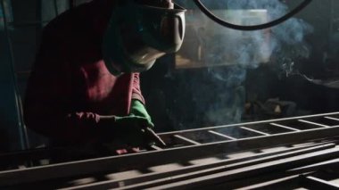 Koruyucu teçhizattaki demirci kaynak makinesini kullanarak merdivenin metal parçalarını eritiyor. Erkek kaynakçı atölyede parlak kıvılcımlar ve duman yaratır