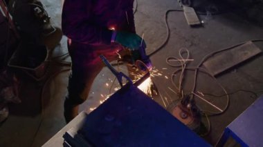 Üniformalı metal işçisi metal bloğu öğütüyor. Masanın önünde duran eldivenli adam dükkanda malzeme ve sabitlenmiş klemple çalışıyor.
