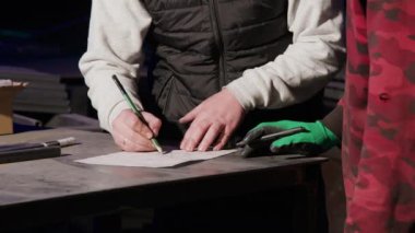 Eldivenli demirci ve gömlekli mühendis masanın üstündeki kağıt çizimlerini düzeltiyor. Metal işçisi nesnenin nihai tasarımında iyileştirmeler önerdi
