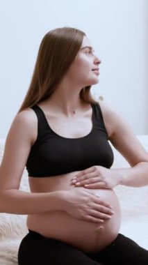 Gülümseyen hamile kadın evdeki koltukta otururken karnını nazikçe okşuyor. Siyah spor sutyenli rahat genç bayan, bebeğin bu anın tadını çıkarmasını bekliyor.