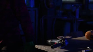 Adam atölyede yürüyor ve masada duran öğütücünün yanından geçiyor. Eldivenli demirci işten sonra geniş atölyeden aletlerle çıkıyor.