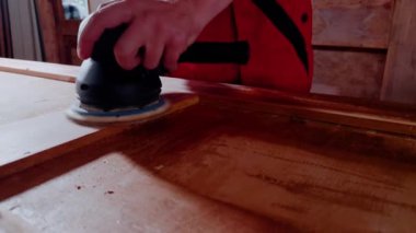 Elinde öğütücü olan ahşap işçisi kapı tahtasında bir cihaz çalıştırıyor. Genç marangoz, boya aşamasından önce kusurları dikkatlice ortadan kaldırarak yüzeyi işliyor.