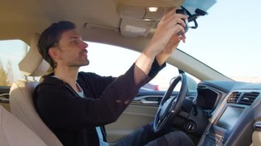 Adam dikiz aynasını araba paylaşımlı araç kuaförüne ayarlıyor ve net bir görüş sağlıyor. Erkek, araba yolculuğuna hazırlanırken son ayarlamaları yapar.