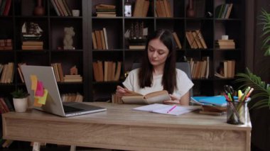 Bayan öğrenci masada kitap okuyor ve edebiyatla dolu büyük bir kitaplığın yanında oturuyor. Genç bir kadın çalışmak için rahat ve aydınlık bir yer seçiyor.