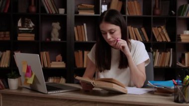 Koyu renk saçlı bayan ders kitaplarını inceliyor. Enstitü öğrencisi kütüphanede teknolojik aygıtlar ve edebiyat kullanılarak sınava hazırlanıyor