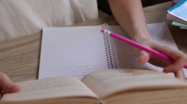 Kadın öğrenci not defterine pembe kalemle sol eliyle notlar yazar ders kitabını bilgi kaynağı olarak kullanır. Kütüphanede sınava hazırlanılıyor.