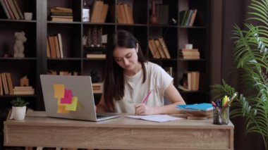 Genç kız öğrenci dizüstü bilgisayara bakarken notlar alır. Solak kadın, kütüphanedeki masada online akademik makaleyi incelerken deftere yazıyor.