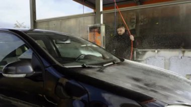 Genç adam arabanın dışını yüksek basınçlı temizleyici hortumla dolduruyor. Adam zaman kazanıyor ve arabasını dışarıda oto yıkamada yıkıyor siyah araç da oto yıkamada.
