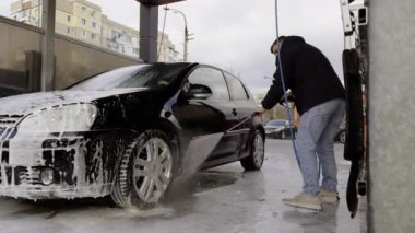 Genç bir adam arabasını yüksek basınçlı su hortumuyla oto yıkamada temizliyor. Adam zaman kazanıyor ve arabasını kendisi yıkıyor. self-servis oto yıkama işi.