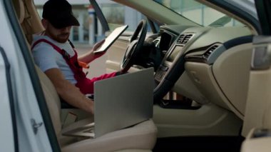 Uzman araba durumunu dizüstü bilgisayar ve tabletle kontrol ediyor. Araçta oturan adam, gösterge panelinde çalışan elektronik bileşenlerin dijital analizini yapar.