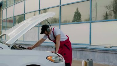 Kulaklıklı tamirci araba teftişi yapan müzik dinler. Teknisyen araç parçalarını analiz eder ve otomobil denetimini tamamlamak için geri adım atar.