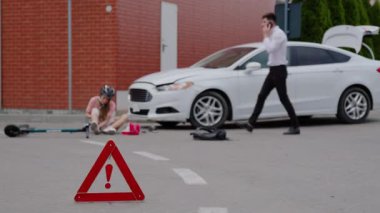 Klasik takım elbiseli bir adam elektrikli scooter kullanan bir kadına çarpmış. Kızgın bir genç adam telefonda konuşuyor ve ambulans, polis ve sigorta şirketini arıyor. Uyarı işareti.