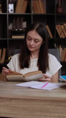 Bayan öğrenci masada kitap okuyor ve edebiyatla dolu büyük bir kitaplığın yanında oturuyor. Genç bir kadın çalışmak için rahat ve aydınlık bir yer seçiyor.