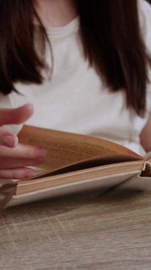 Açık renkli tişörtlü bir kadın kütüphanede masada kitap boyunca oturuyor. Kendini kaptırmış okuyucu sayfalarca hikayeye gömüldü.