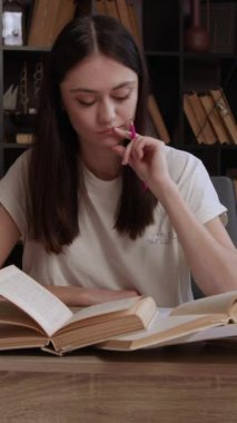 Genç esmer öğrenci not defterine not yazmak için kitap topluyor. Üniversite kütüphanesinin sessiz ortamında sınav hazırlığı