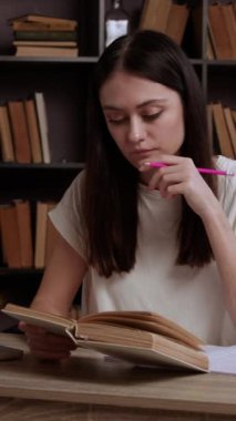 Koyu renk saçlı bayan ders kitaplarını inceliyor. Enstitü öğrencisi kütüphanede teknolojik aygıtlar ve edebiyat kullanılarak sınava hazırlanıyor