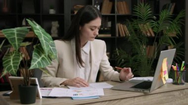 Klasik takım elbiseli öğrenci stajyer bir ofis masasında oturuyor. Kadın birkaç şirketin istatistik ve grafiklerini analiz ediyor ve birkaç büyüme stratejisi oluşturmayı düşünüyor. İş geliştirme şirketi.