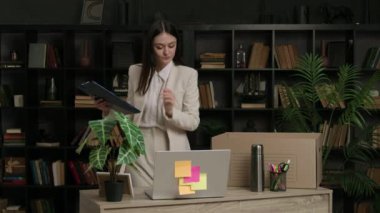 Düşünceli ve mutlu bir kadın kişisel eşyalarını toplayıp karton bir kutuya koyar. Neşeli kadın kağıtların üzerinde eşyaların yeni ofise taşınması gerektiğini yazıyor.