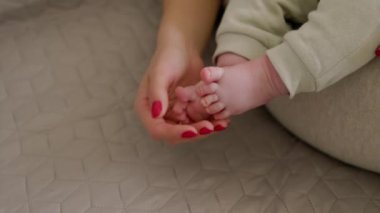Bir annenin elinin, bebeğin küçük ayakları ve parmaklarıyla nazikçe oynadığı yakın plan görüntüsü. Bebek annesinin kollarında oturuyor ve nazikçe ayaklarıyla oynuyor. Parlak, rahat bir ev. Güzel annelik