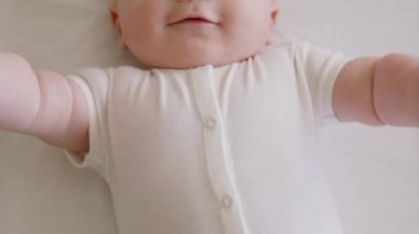 Gülümseyen neşeli ve aktif bir bebeğin yakın plan görüntüsü. Beyaz elbiseli bebek mutlu bir şekilde gülümseyip kollarını sallıyor ve bacaklarını kaldırıyor. Yeni doğmuş bir çocuğun masum gülümsemesi.
