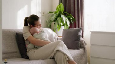 Genç ve güzel bir anne, parlak bir odada bir kanepede oturup bebeğini kollarında tutuyor. Genç anne yeni doğmuş bebeğiyle her anın tadını çıkarıyor. Huzurlu ve güzel annelik.