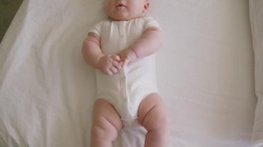 Beyaz elbiseli tombul bir çocuk, parlak bir odada, yatak odasında uzanmış ellerini ve ayaklarını tek başına oynatıyor. Neşeli ve aktif küçük çocuk. Sağlıklı, gelişmiş bir çocuk. Yukarıdan görüntü