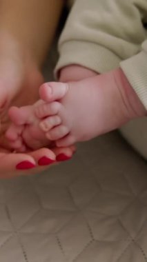 Bir annenin elinin, bebeğin küçük ayakları ve parmaklarıyla nazikçe oynadığı yakın plan görüntüsü. Bebek annesinin kollarında oturuyor ve nazikçe ayaklarıyla oynuyor. Parlak, rahat bir ev. Güzel annelik